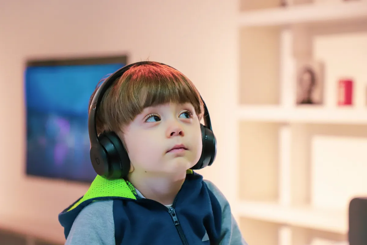 Young child wearing black headphones and a blue jacket with a green hood looking slightly upward indoors.
