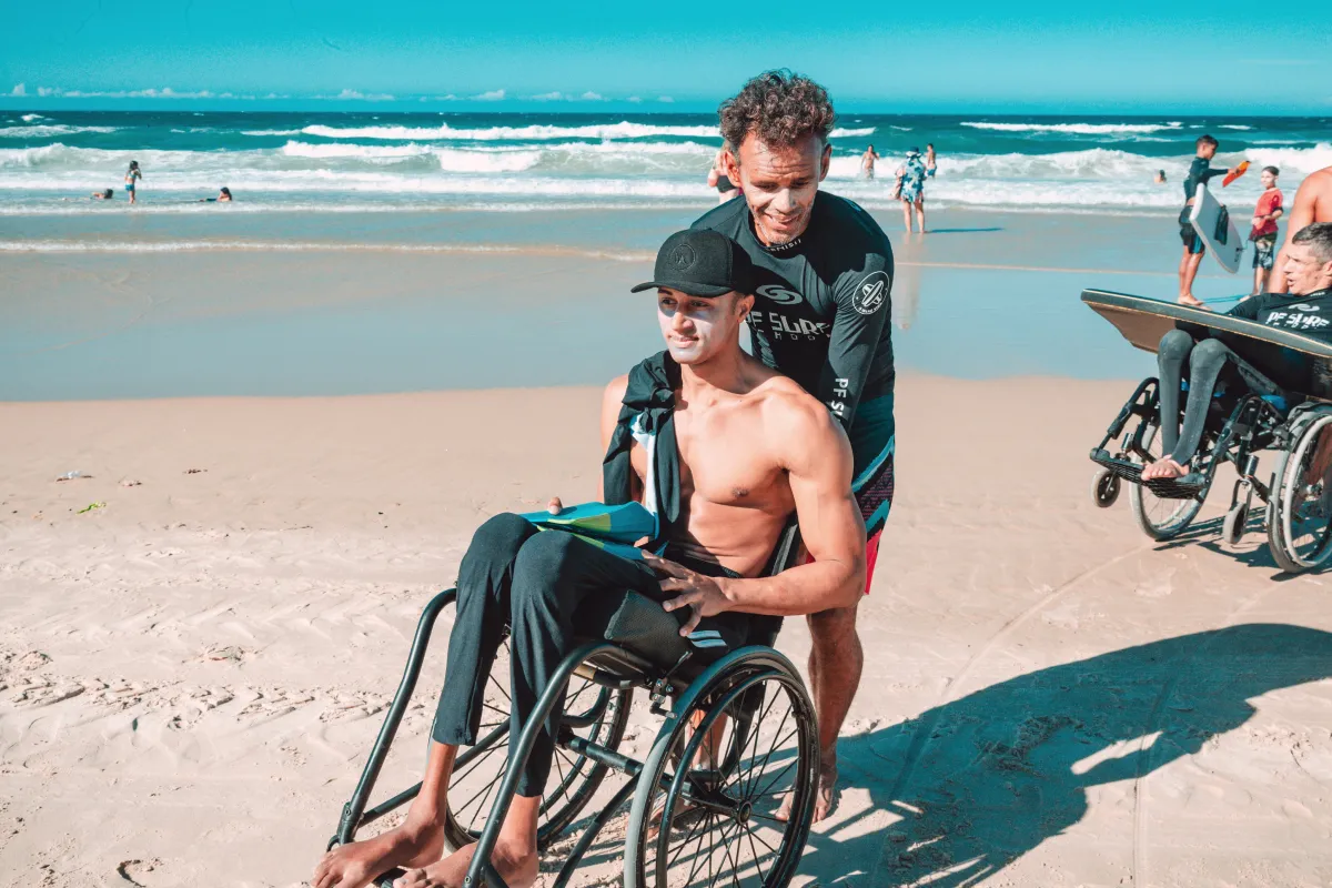 Man in black wetsuit assisting shirtless man in wheelchair on a sandy beach with surfers and ocean waves in the background.