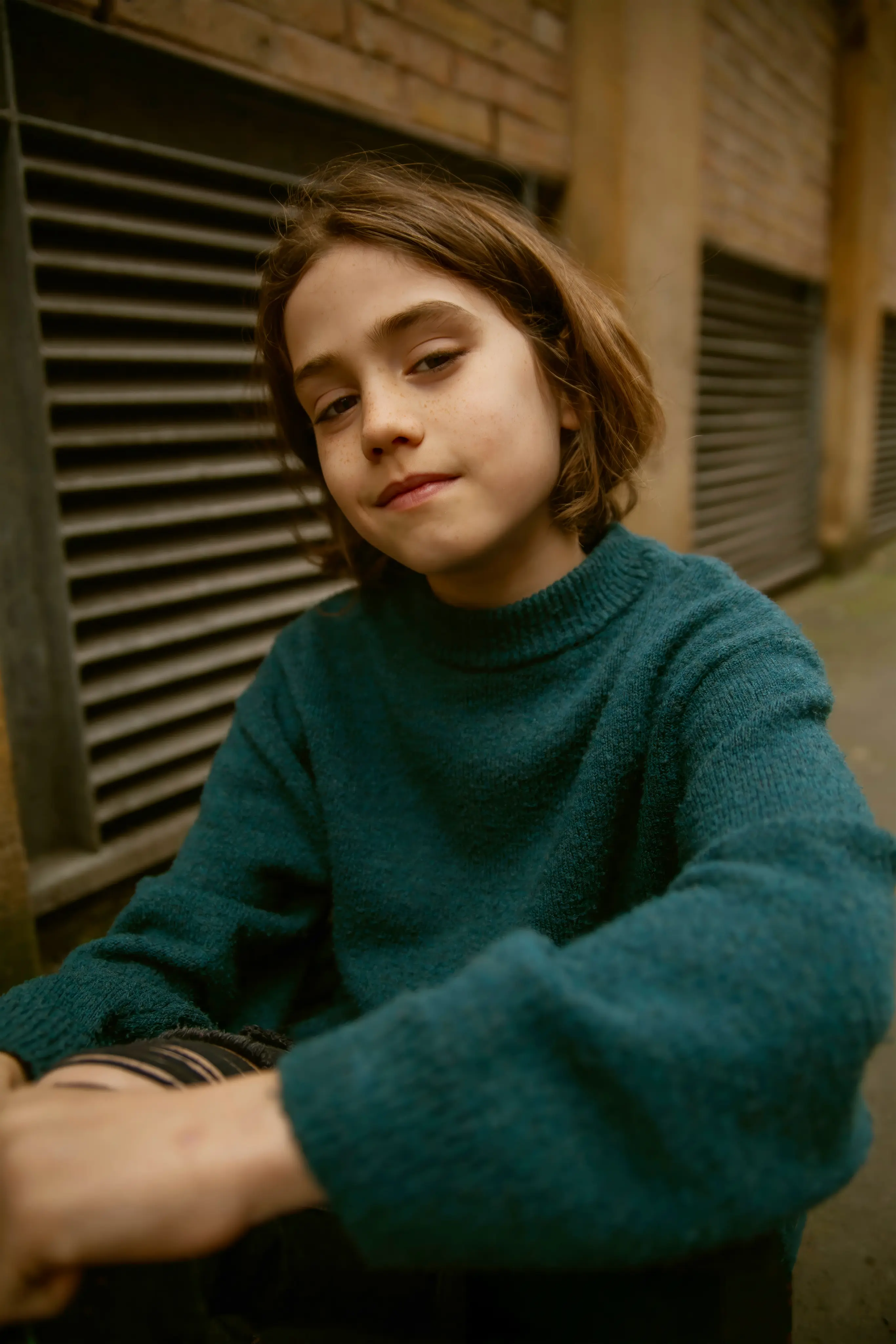 Young person with short brown hair wearing a teal sweater sitting outdoors near a brick wall with vents.