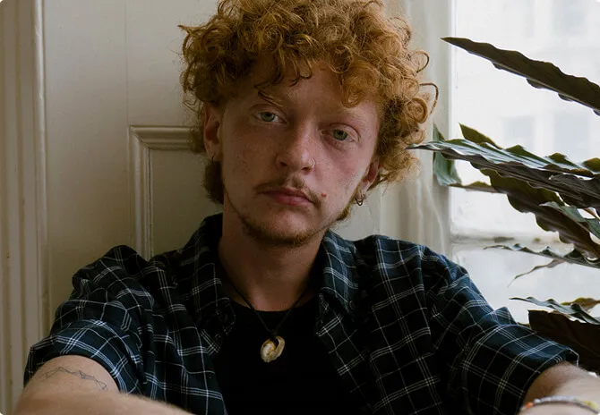 A man with red curly hair sitting in front of a potted plant.