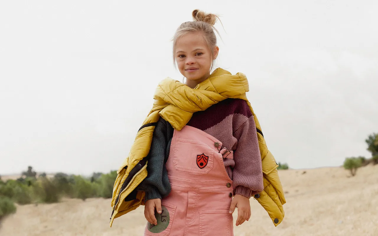 Smiling young girl with blonde hair in a bun wearing a colorful sweater, pink overalls, and a yellow jacket draped over her shoulders standing outdoors.