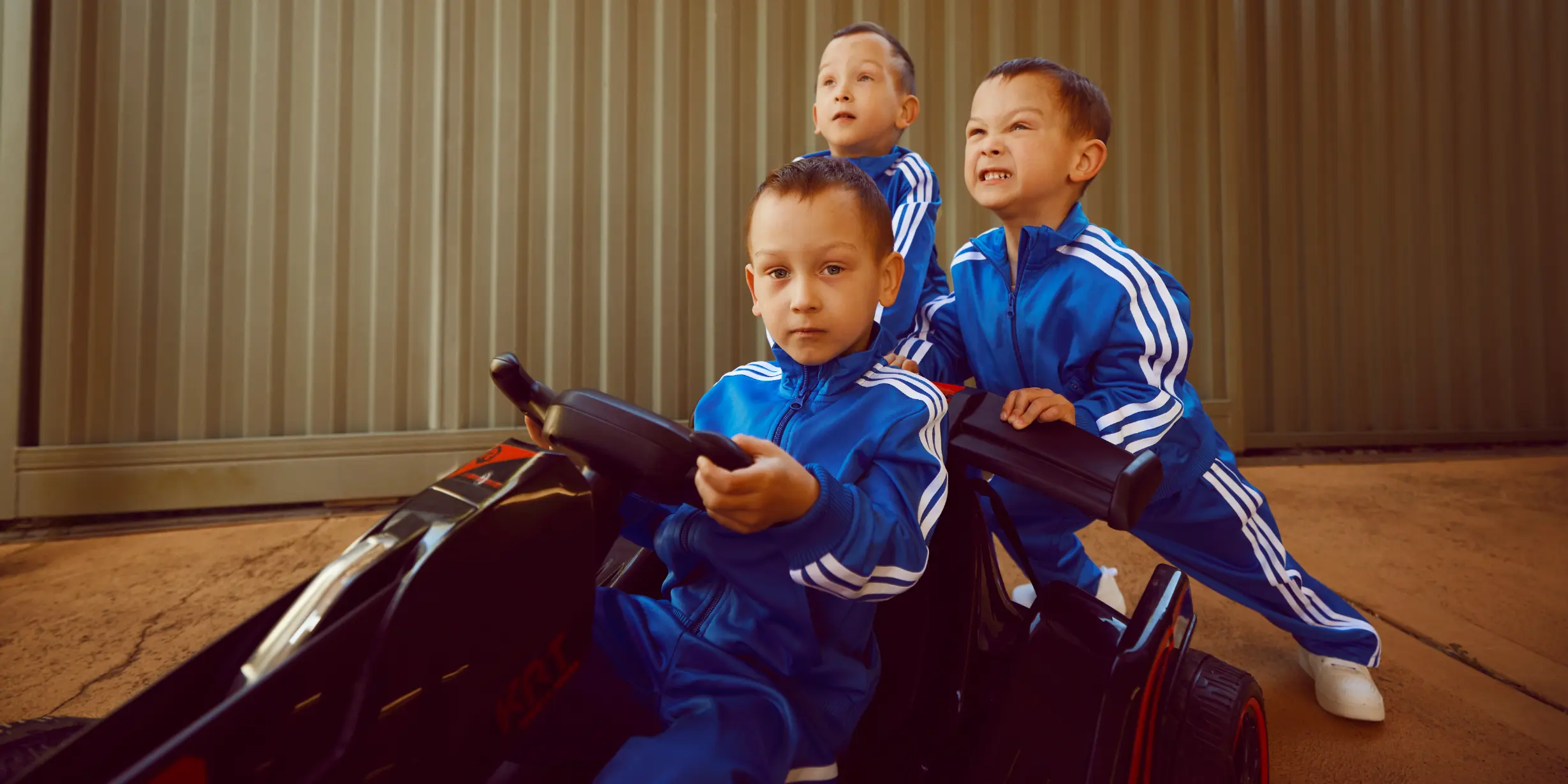 Three boys in matching blue tracksuits with white stripes playing with a black toy car, one boy driving and two standing behind.