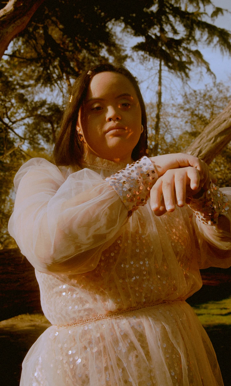 Person outdoors in fancy sheer dress with sequins, posing with hands raised in sunlight.