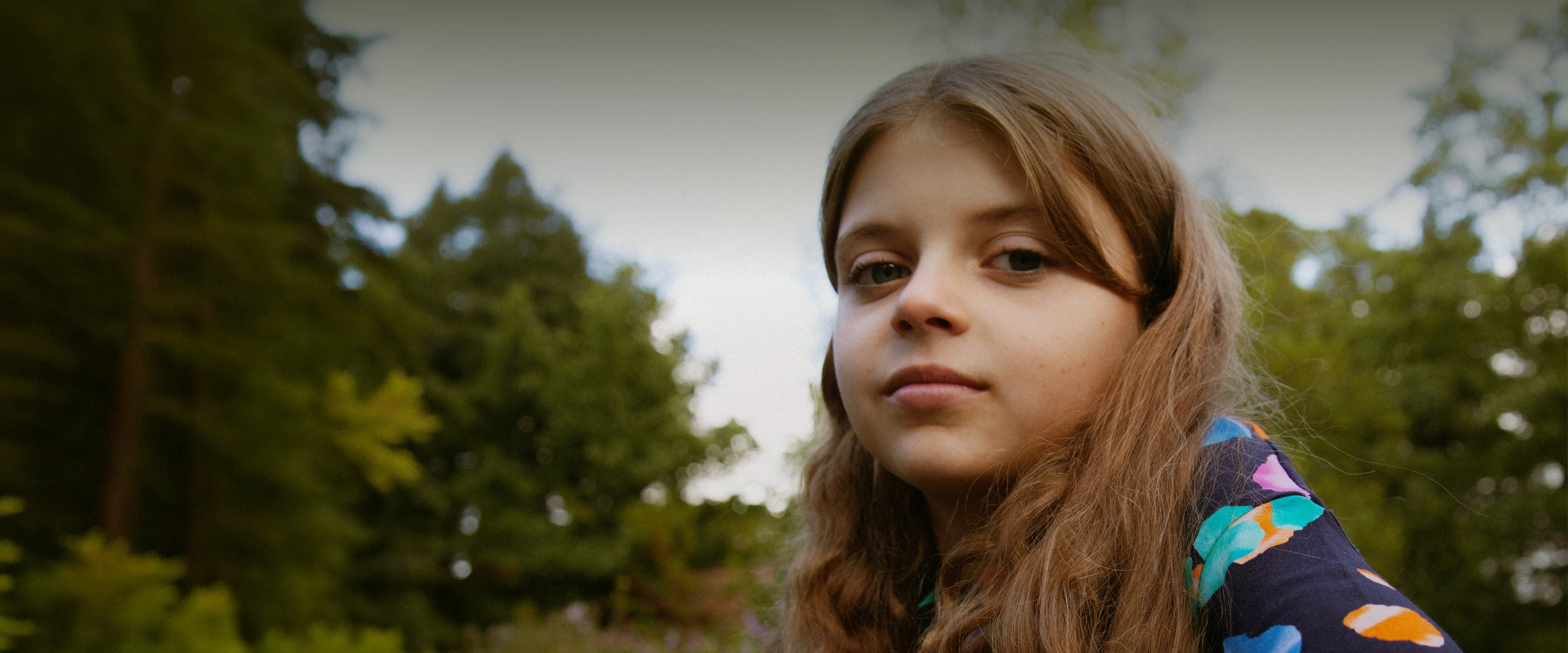 Close-up of a girl with long hair outdoors with blurred green trees in the background.