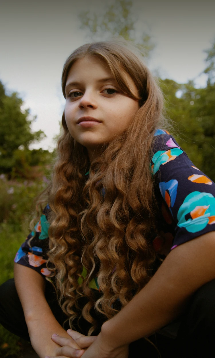 Girl with long curly hair wearing a colorful shirt sitting outside in a natural setting.