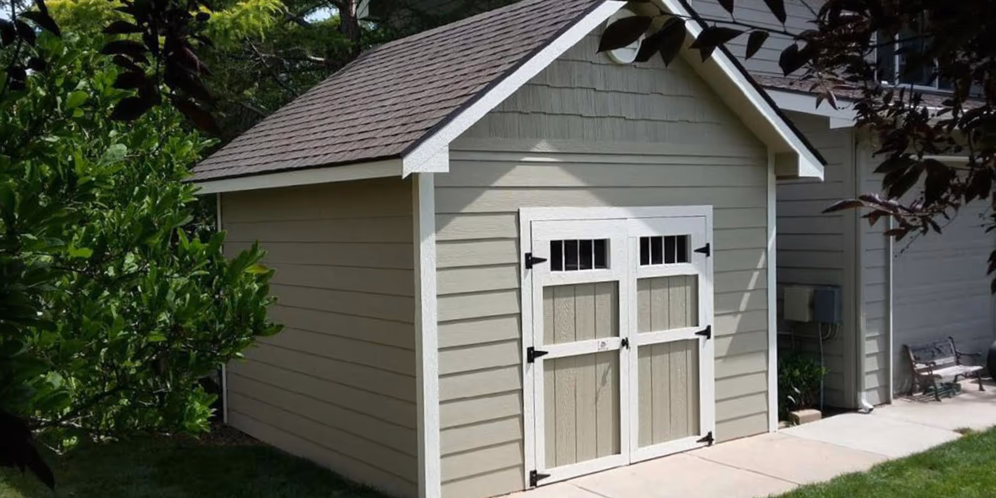 Beige outdoor storage shed with white trim, double doors with black hinges, and a shingled gable roof beside a house and greenery.