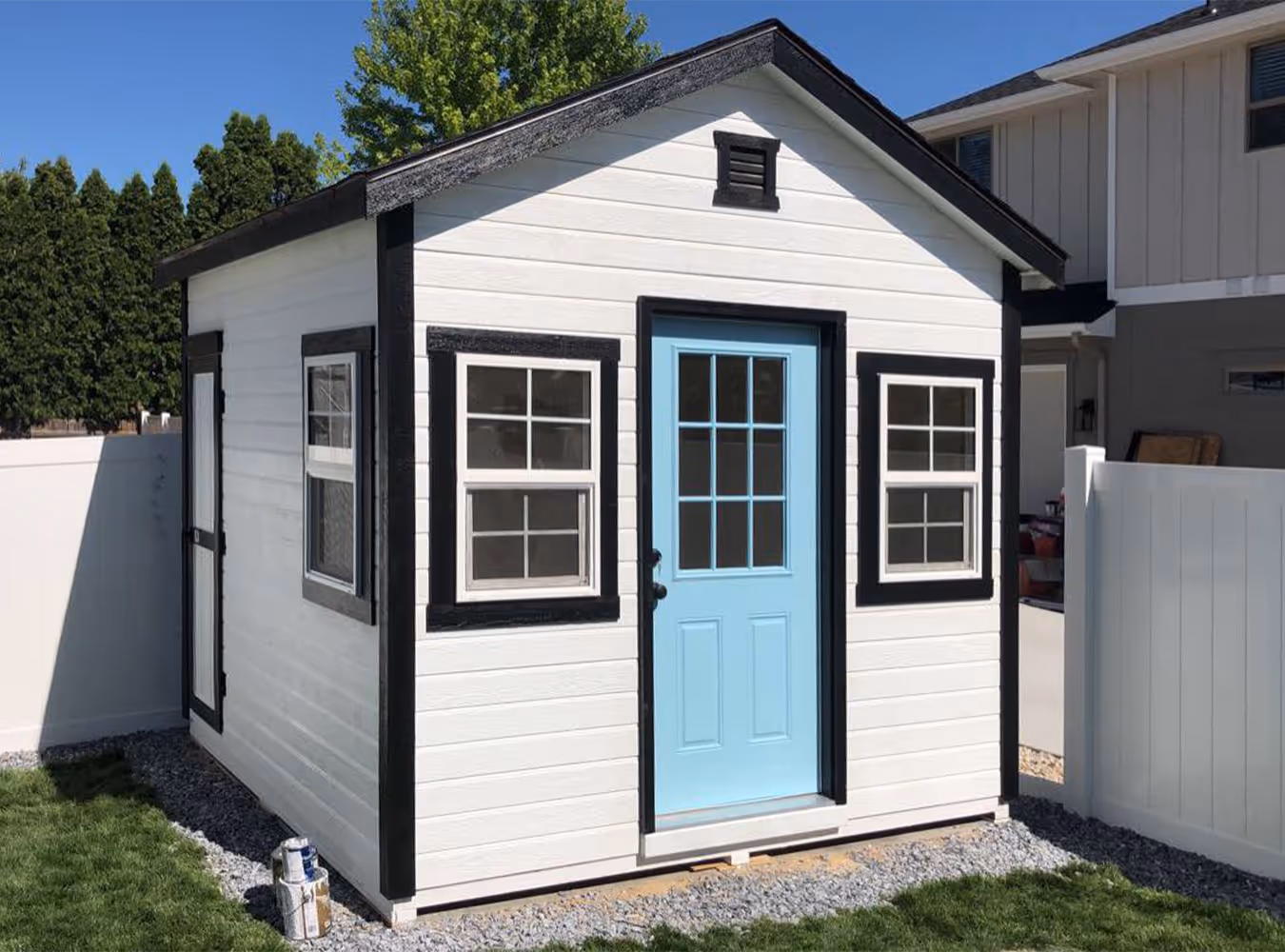Small white backyard shed with black trim and a light blue door, surrounded by grass and gravel.
