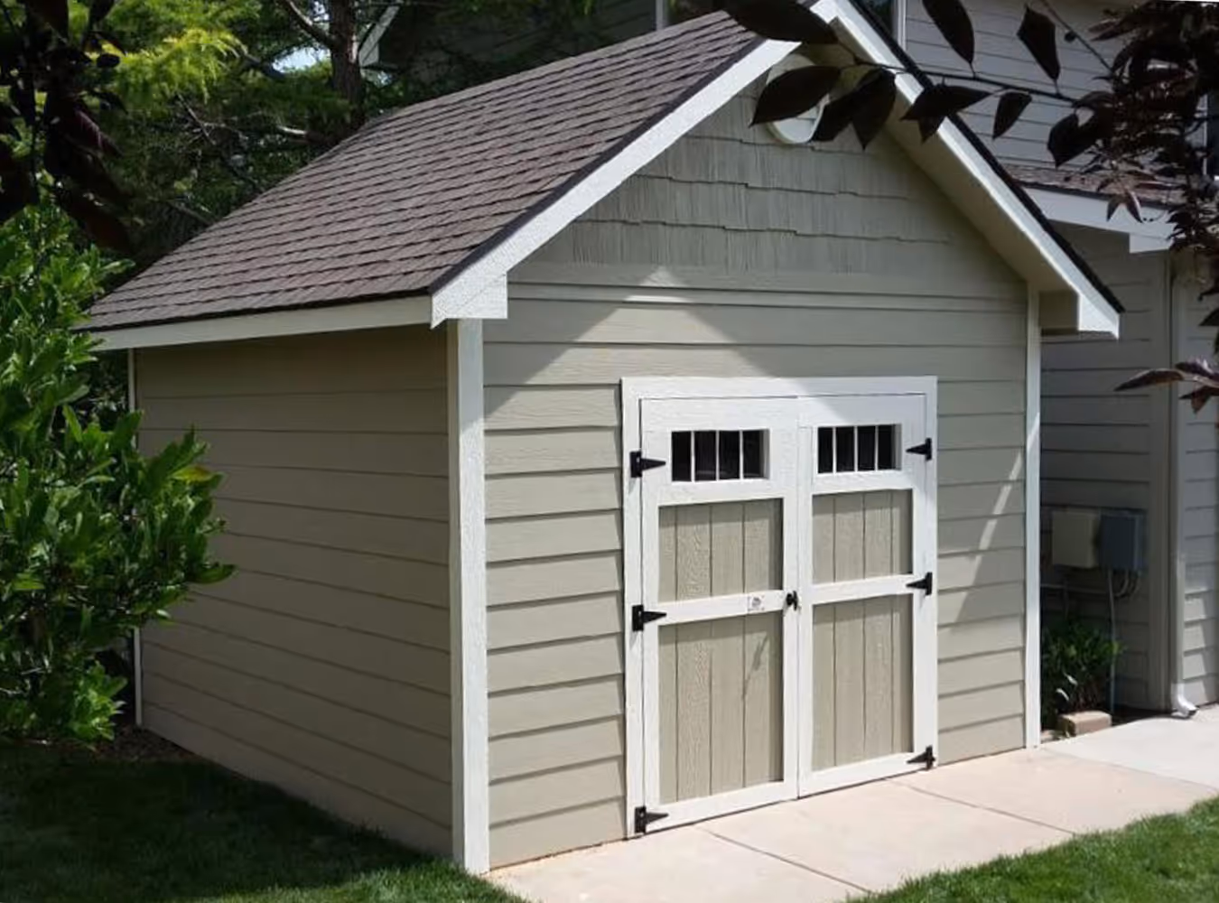 Small beige garden shed with white trim and double doors featuring black hinges and small rectangular windows at the top.