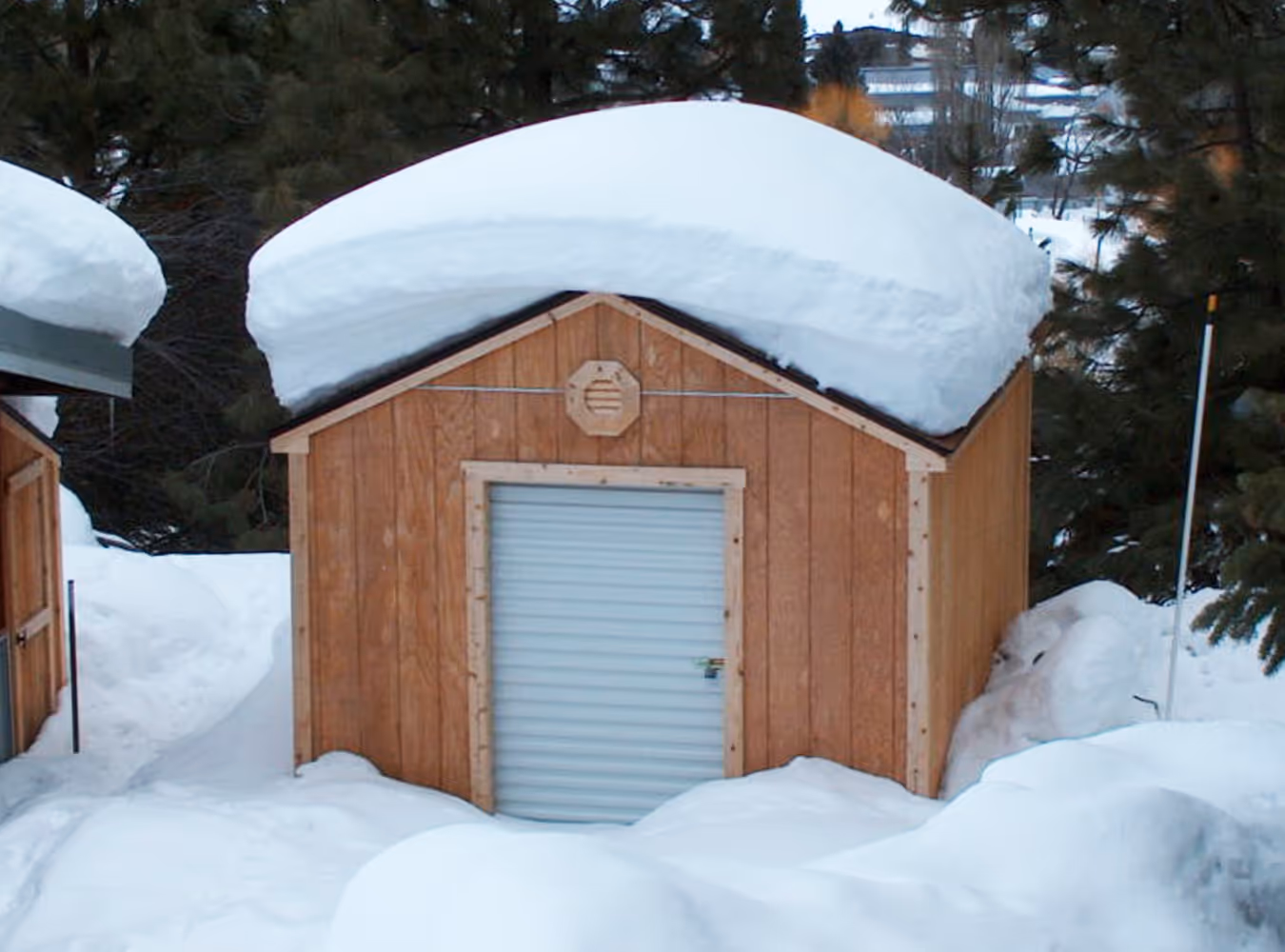 Small wooden shed with a snow-covered roof and a locked metal door, surrounded by snow and pine trees.