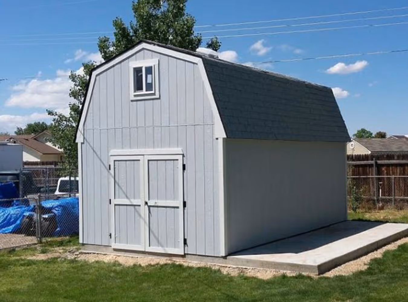 Gray barn-style shed with double doors and an upper window on a grassy yard under a blue sky.