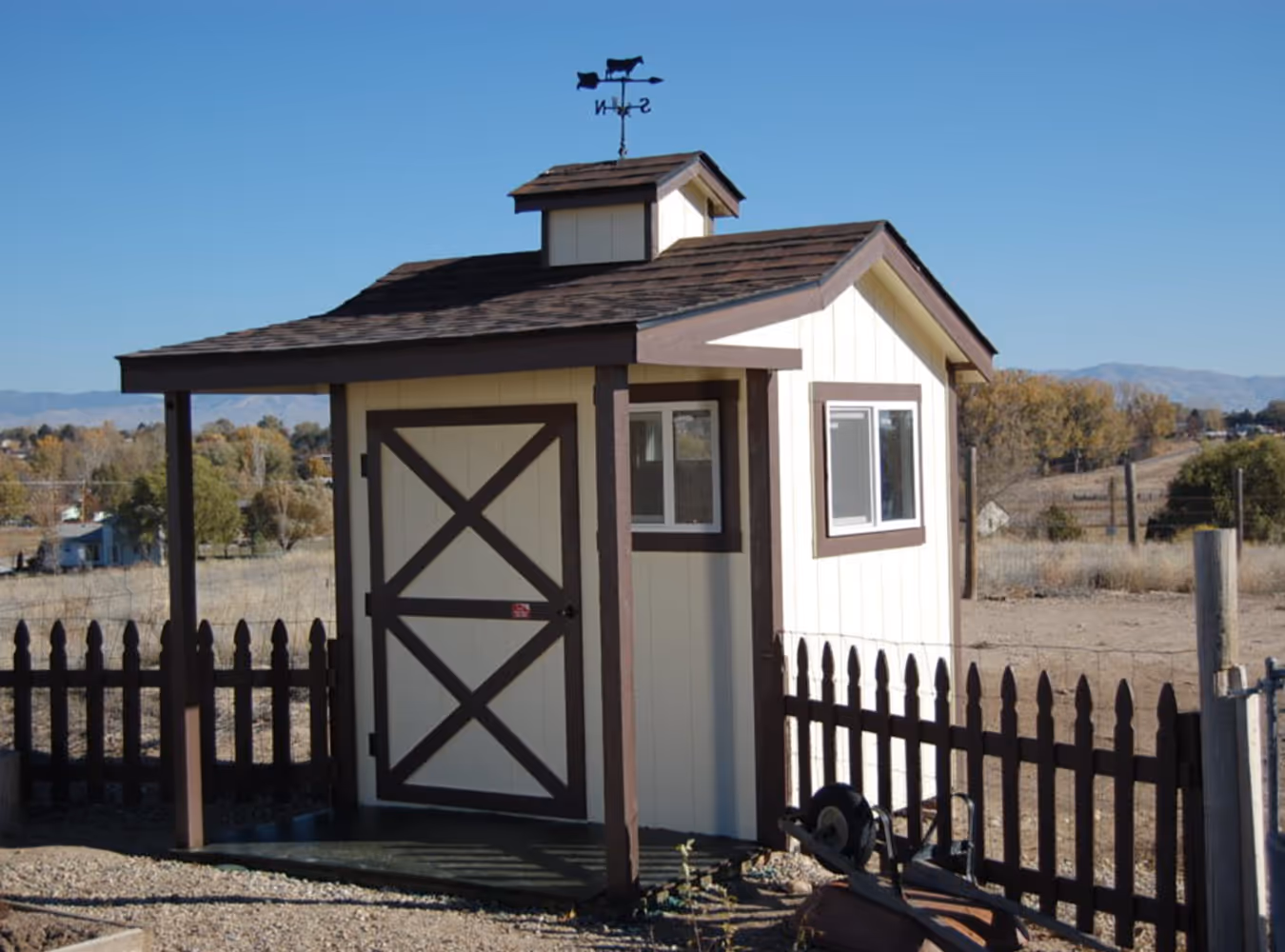 Small beige shed with brown trim, a weathervane on the roof, and a brown picket fence surrounding it in a rural area under clear blue sky.
