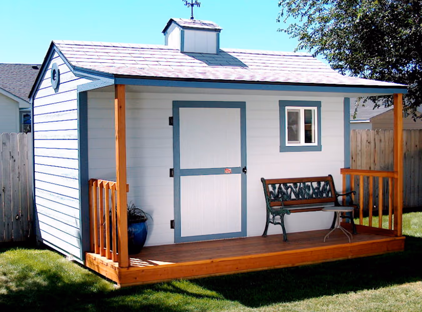 Small white backyard shed with blue trim, a wooden porch, bench, and potted plant under clear blue sky.