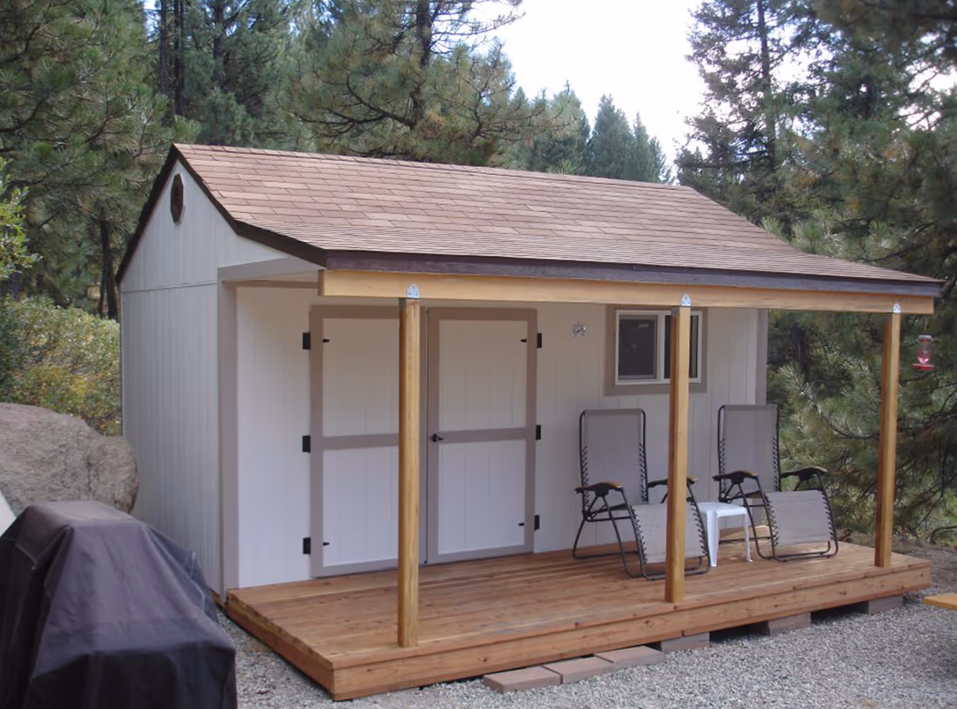 Small white wooden shed with a brown shingled roof, a covered wooden porch with two chairs and a small table, surrounded by trees.
