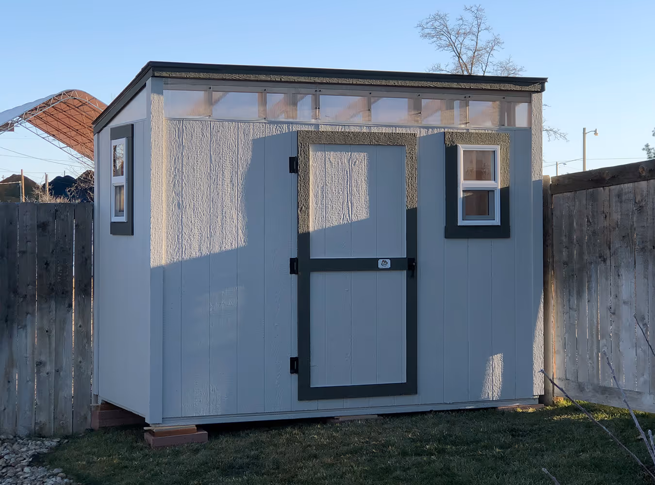 Gray garden shed with a slanted roof, a central door, and two windows on the sides, situated on grass between wooden fences.