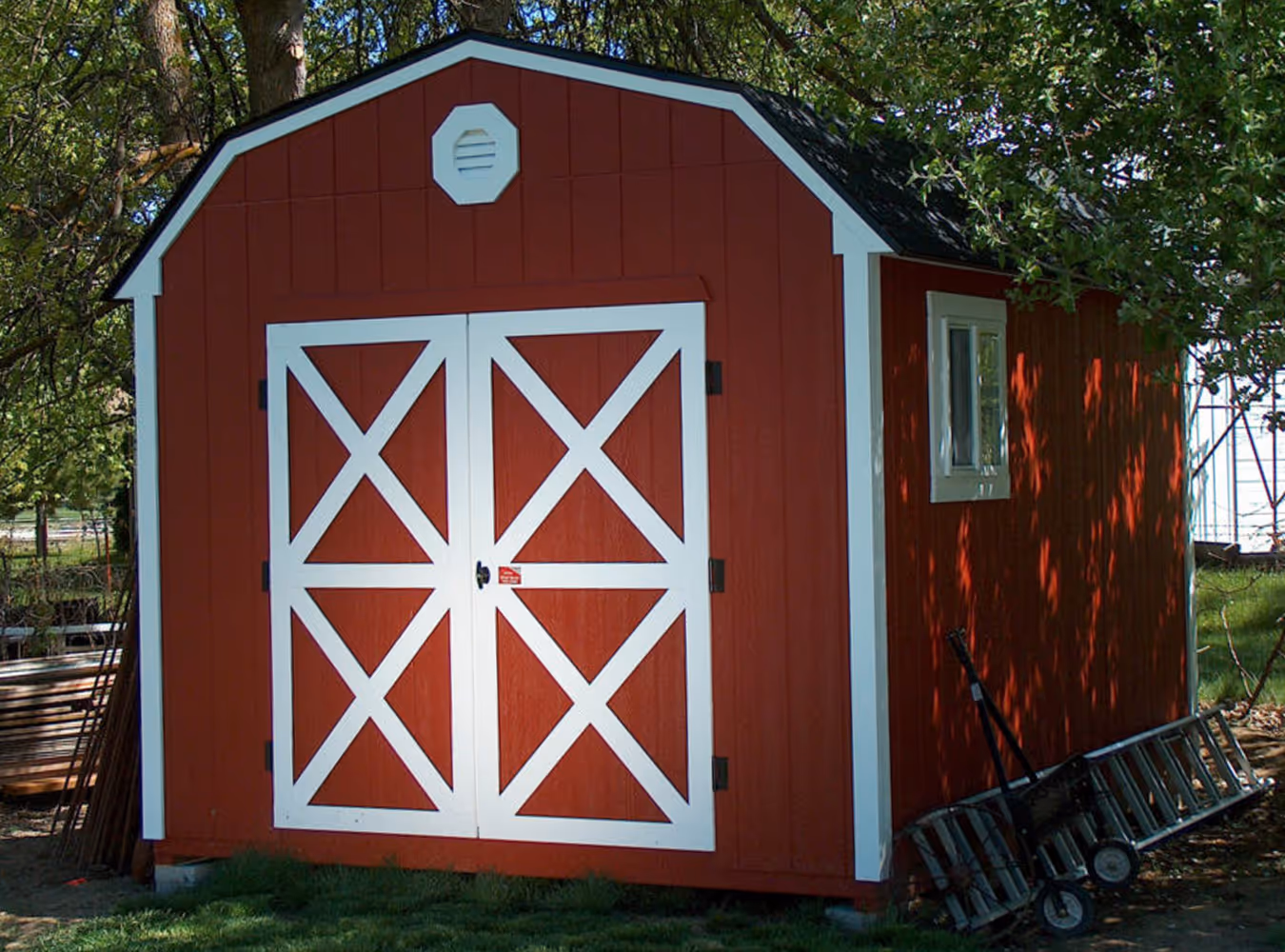 Red barn with white trim and double barn doors, surrounded by green grass and trees, with a ladder and hand truck leaning against the side.