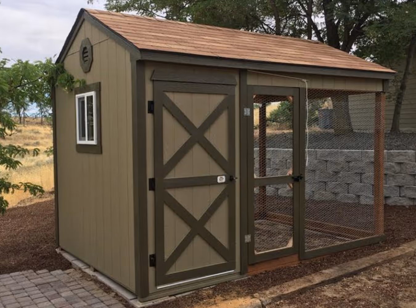 Outdoor chicken coop with a solid wooden door and adjacent wire mesh enclosure, set on a paved and mulched ground.