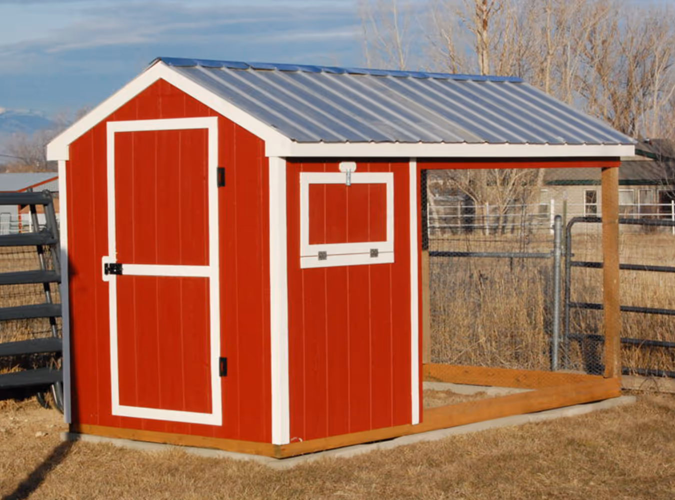 Red wooden chicken coop with white trim and a metal roof, set on a grassy area with a fenced background.