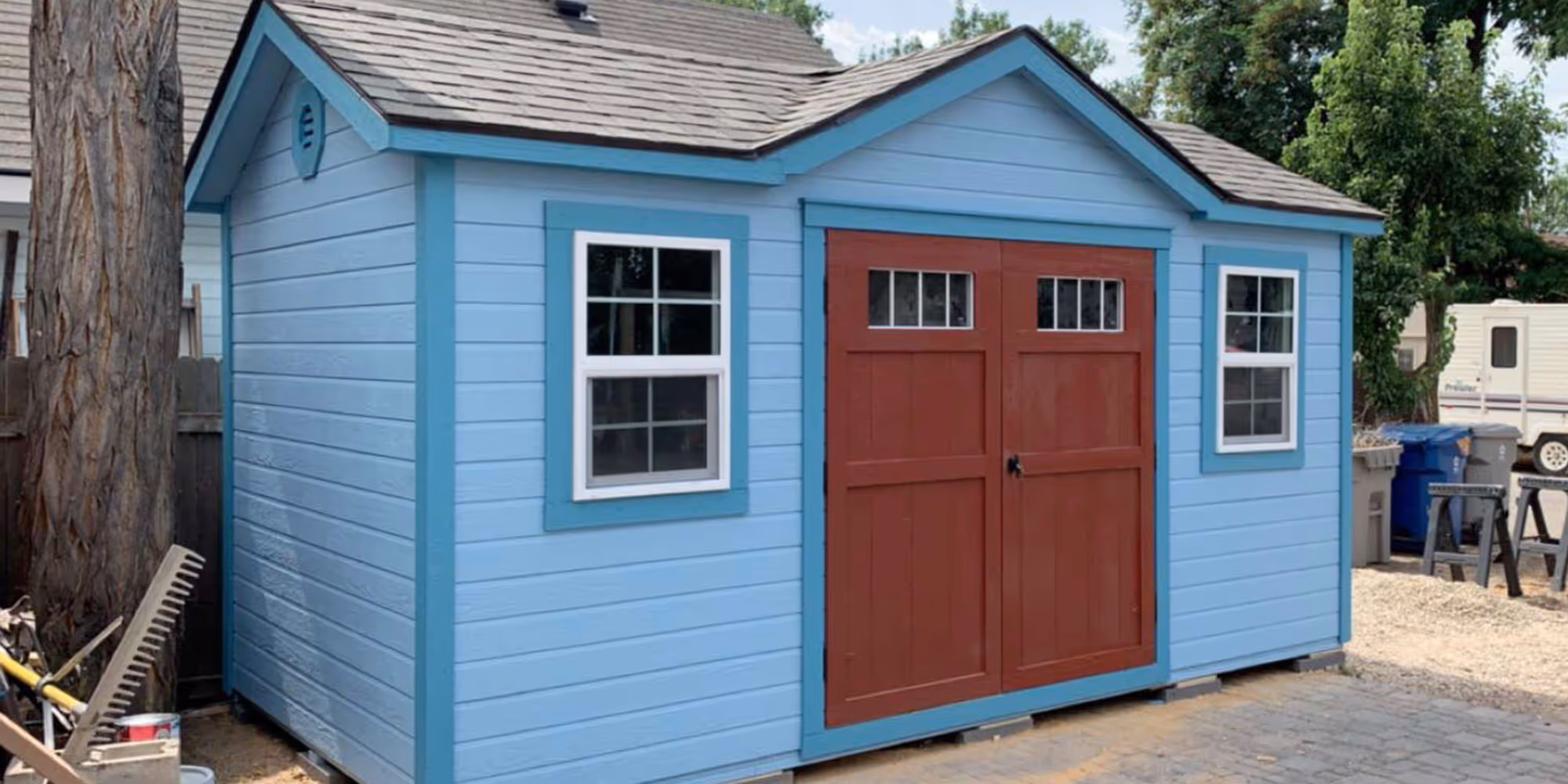 Light blue garden shed with bright blue trim and double reddish-brown doors, two white-framed windows, set on a paved area with trees and waste bins nearby.