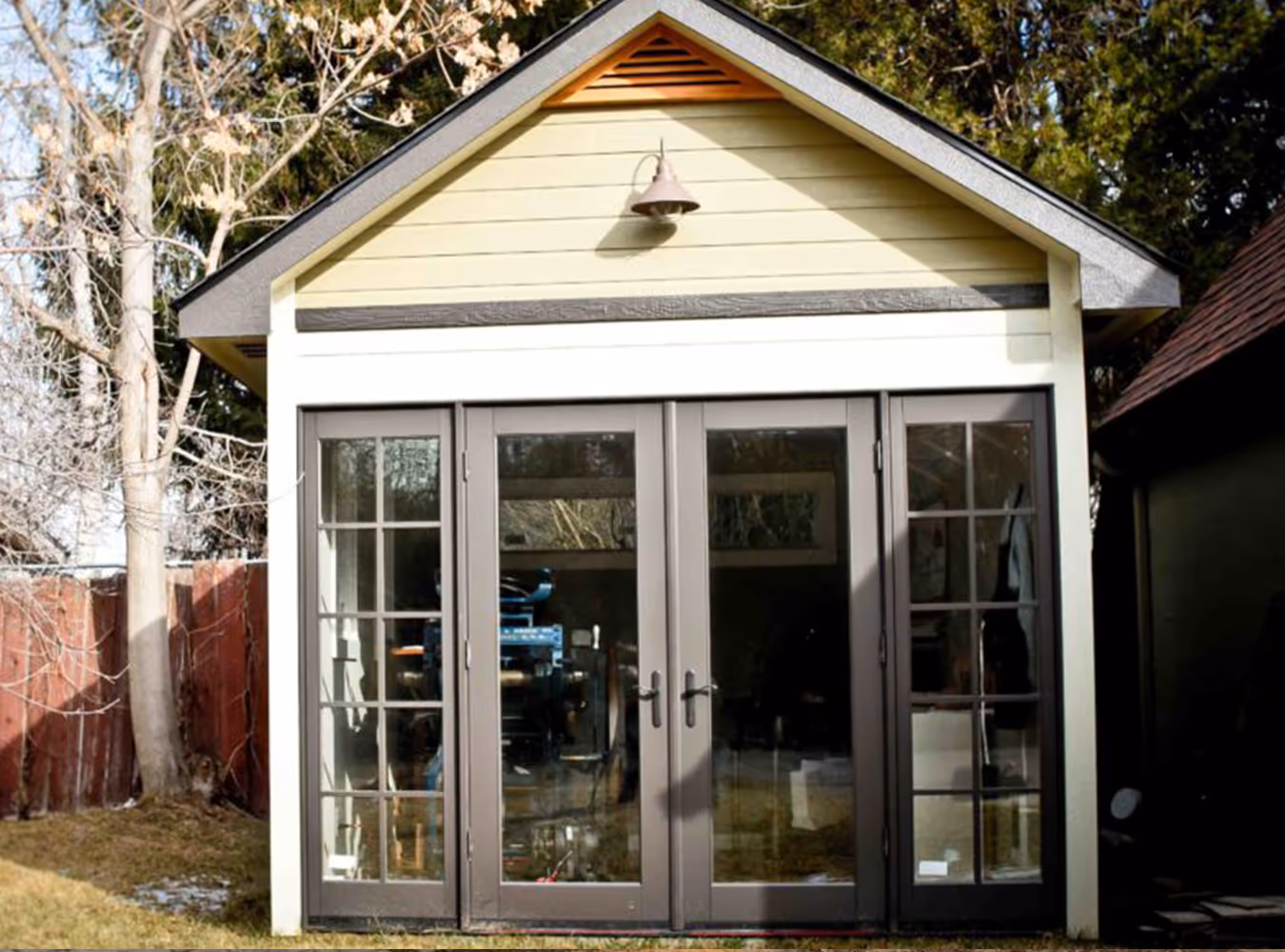 Small backyard shed with beige siding and large dark-framed glass doors under a gable roof.