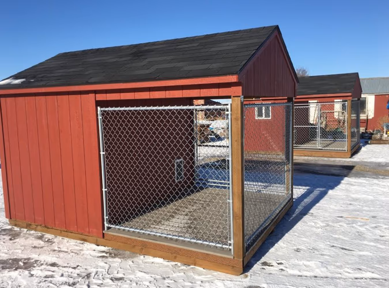 Red wooden dog kennel with black shingled roof and chain-link fenced run on snowy ground under clear blue sky.