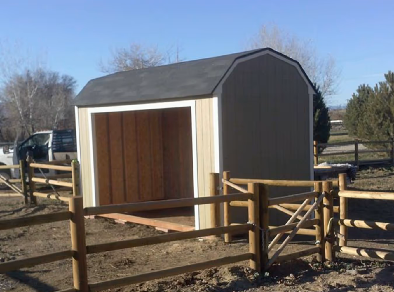 Small open-front wooden shed with a gray roof inside a fenced area on bare soil under a clear sky.