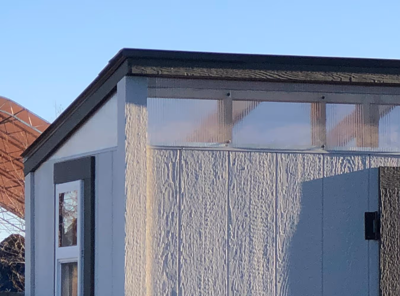 Close-up of a light-colored building corner with textured siding and a small window near the top under clear blue sky.