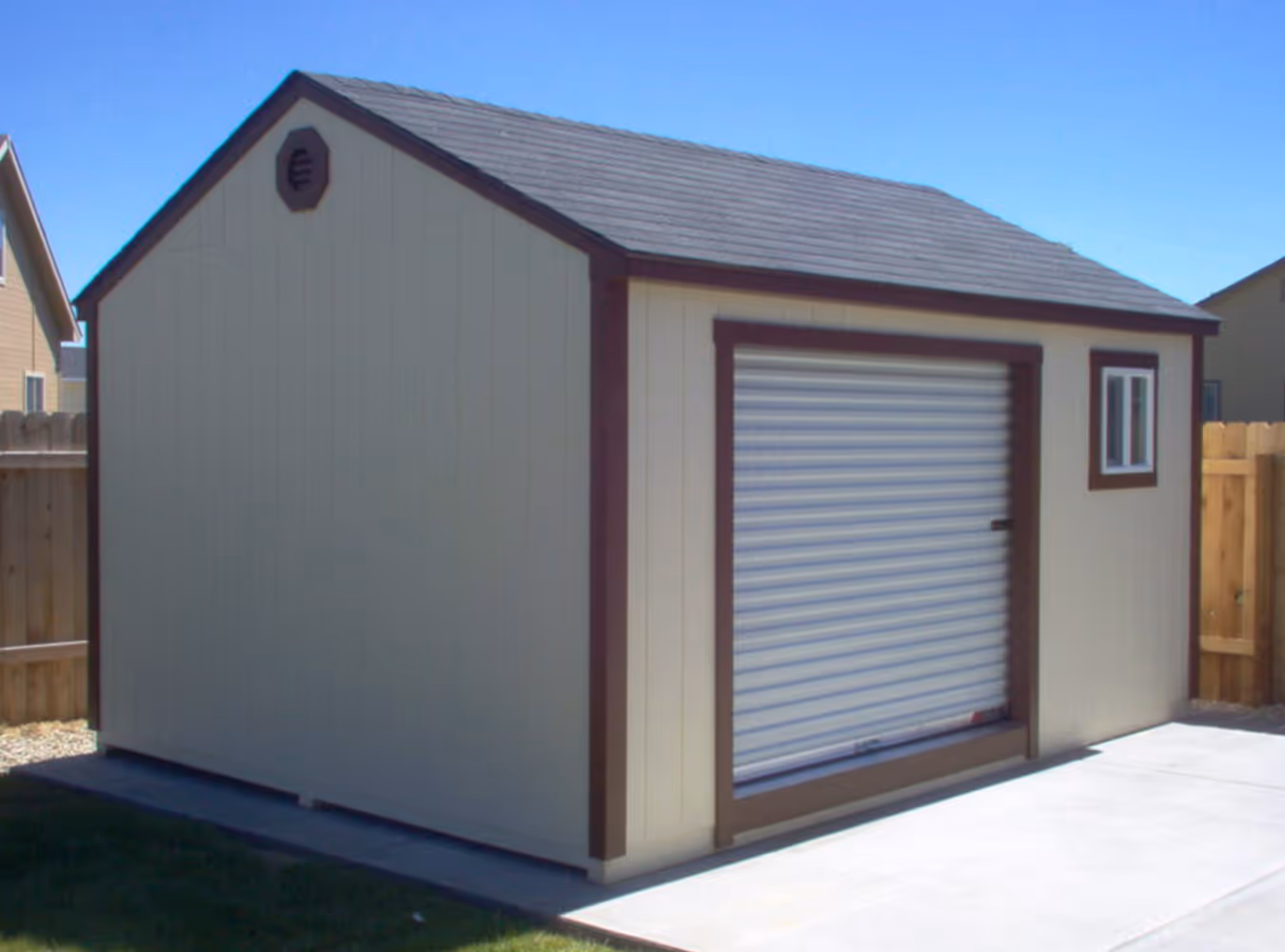 Beige storage shed with brown trim, a closed roll-up door, and a small window, situated on a concrete pad next to a wooden fence.