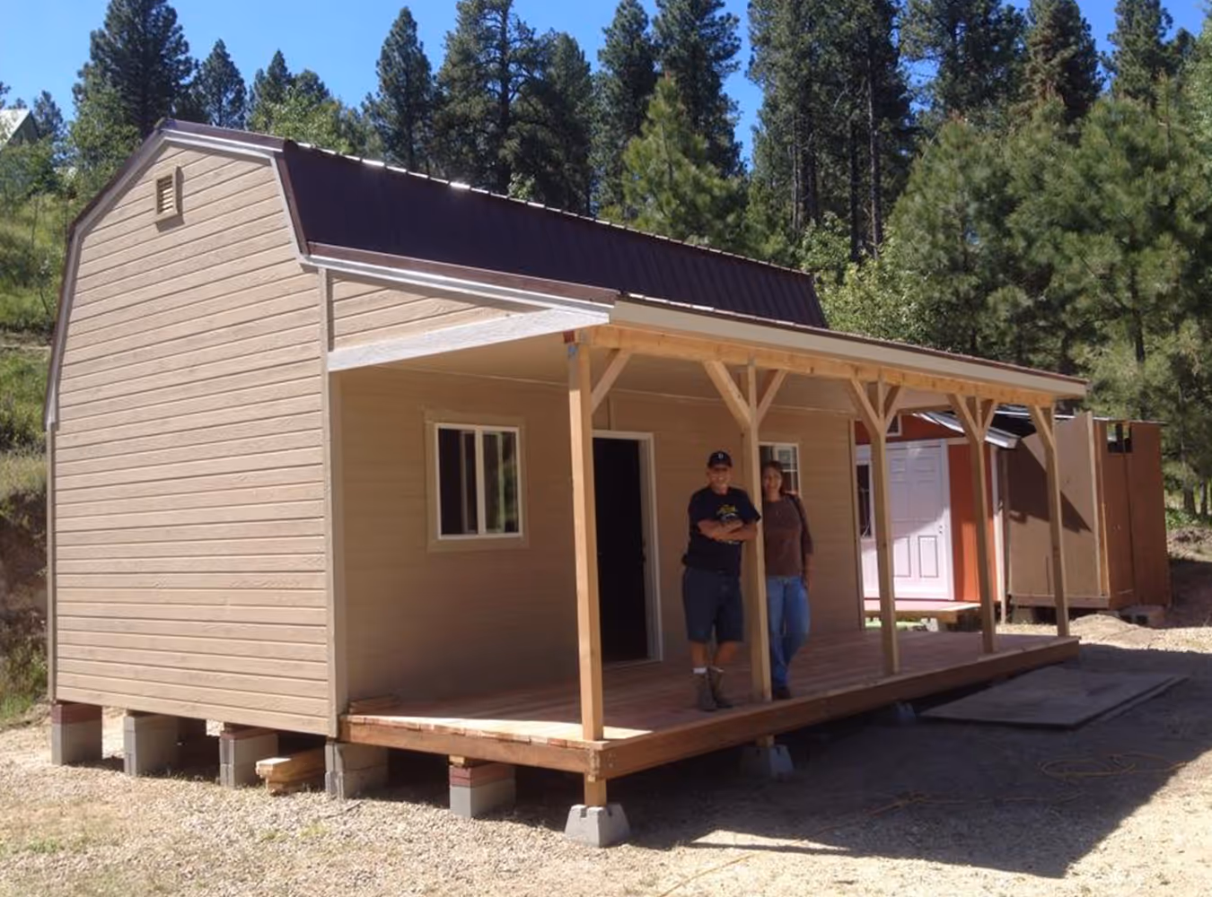 Two people standing on the wooden porch of a small tan cabin with a brown metal roof, surrounded by pine trees.
