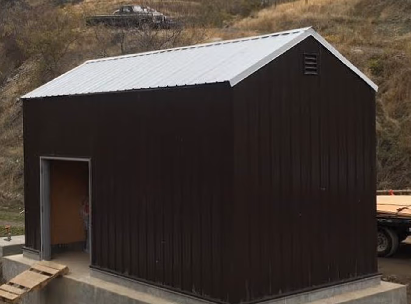 Small dark brown metal shed with a corrugated silver roof on a concrete foundation and an open doorway.