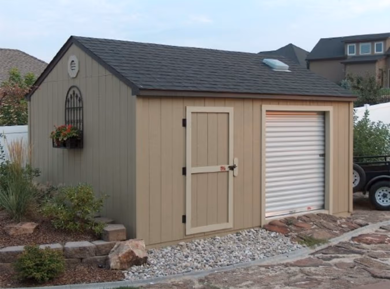 Beige garden shed with a small door, a white roll-up garage door, a dark shingled roof, and a window box with flowers on the side.