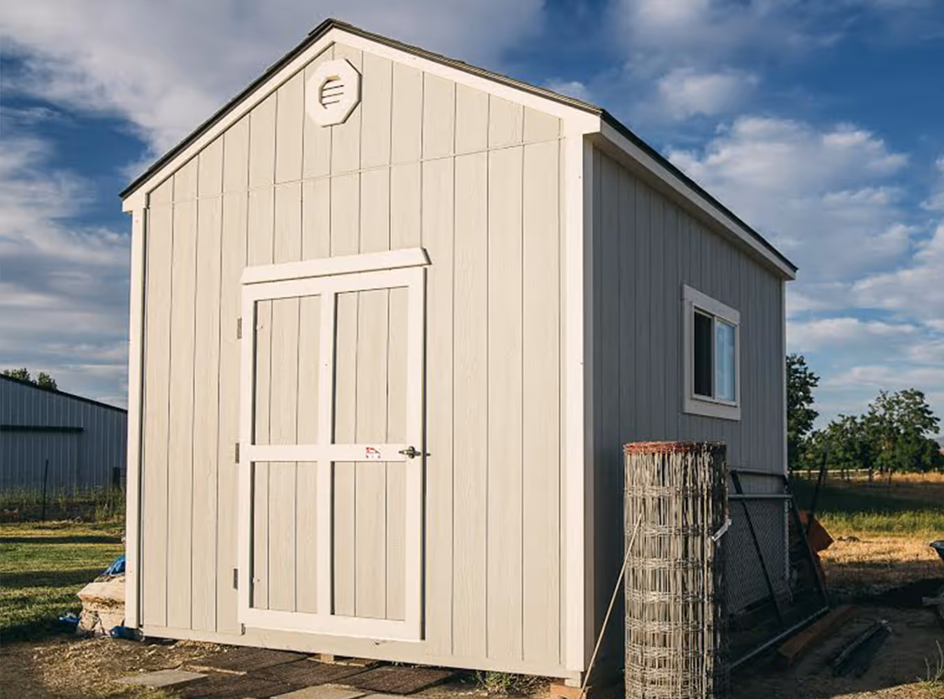 Light gray wooden storage shed with double doors and small window, under a partly cloudy blue sky.