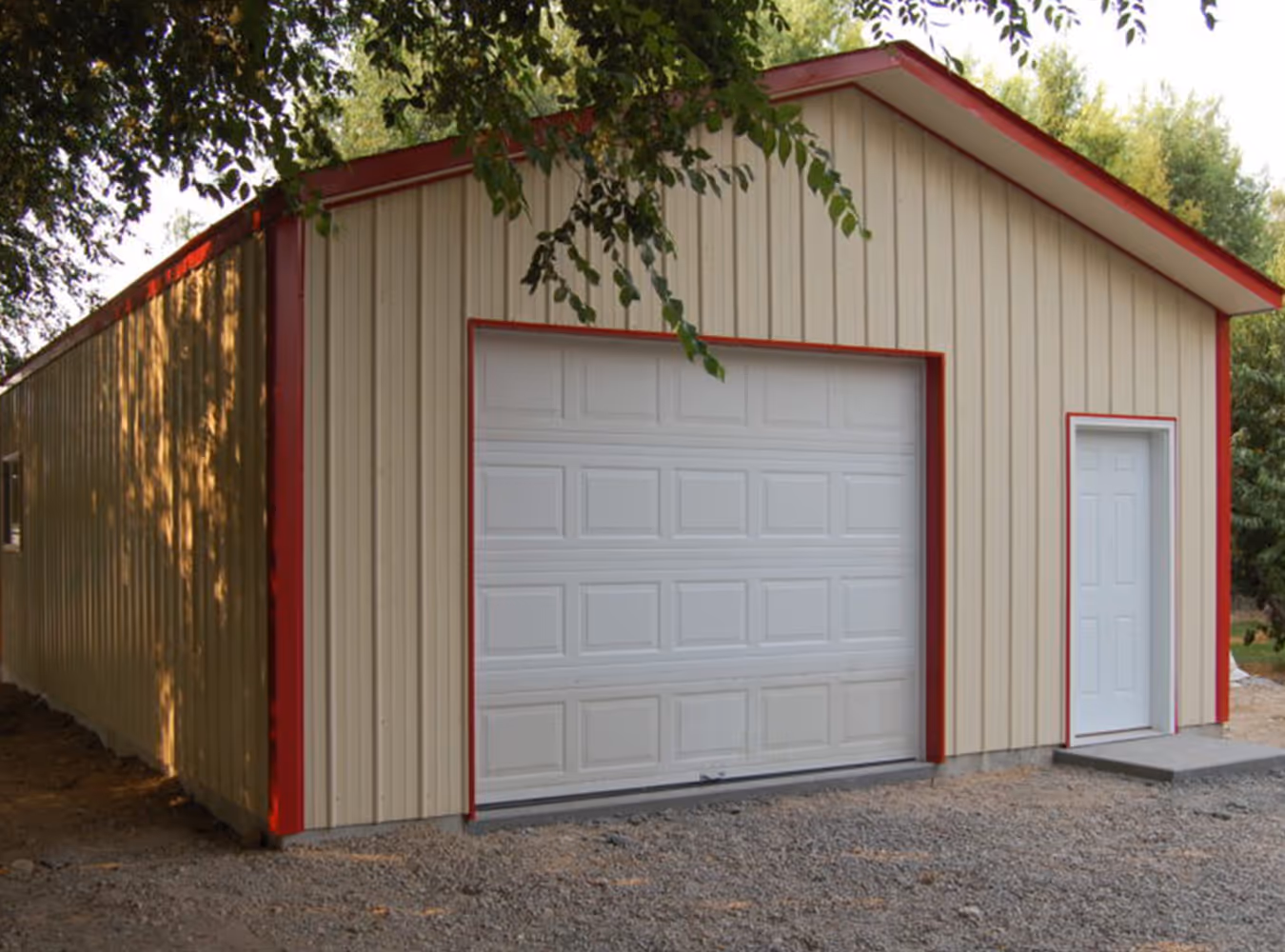 Metal garage building with beige siding, red trim, closed white garage door, and a side entrance door on gravel ground.
