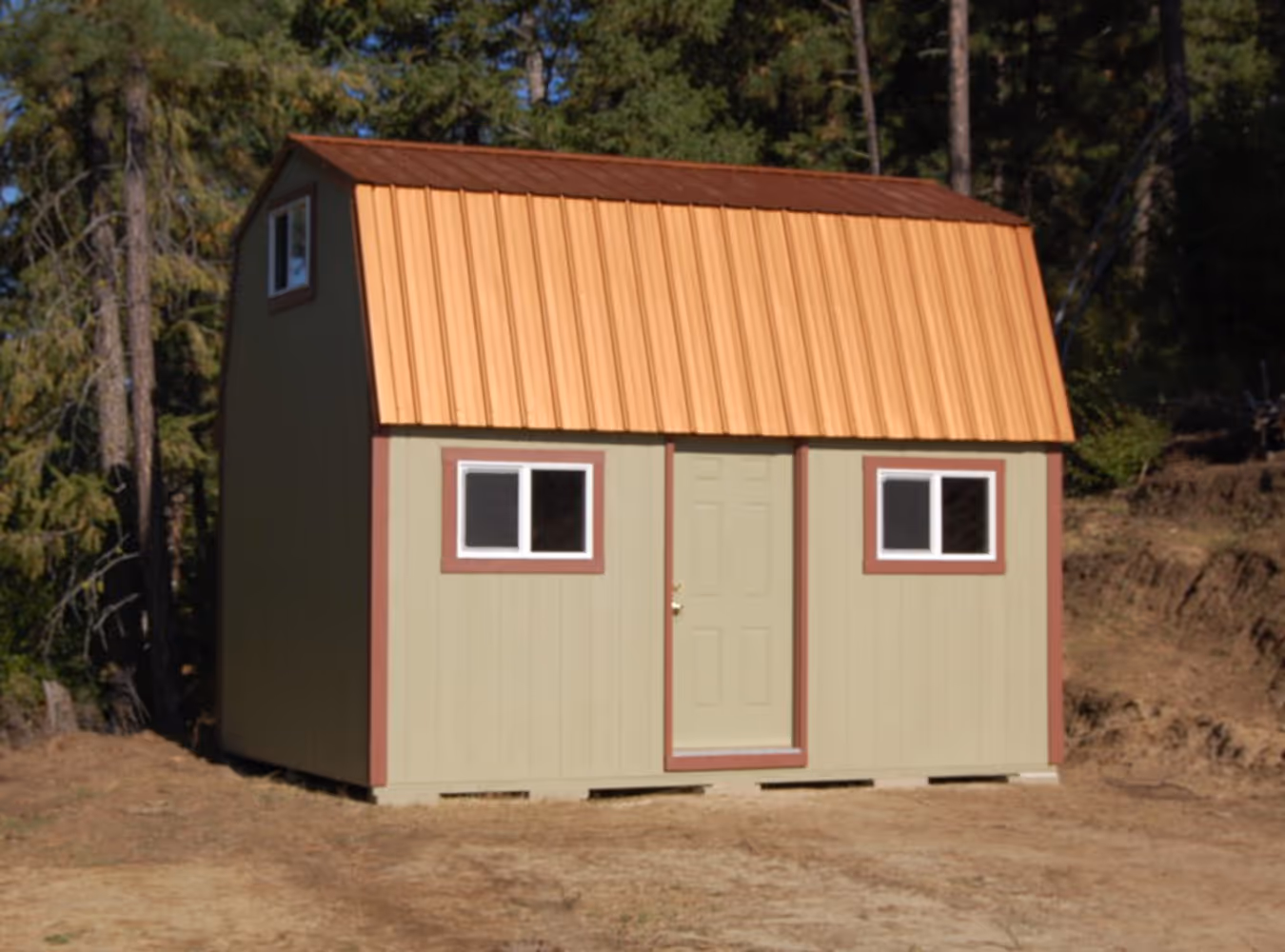 Small barn-style shed with tan walls, a rust-colored metal roof, two windows, and a centered door, set outdoors near pine trees.