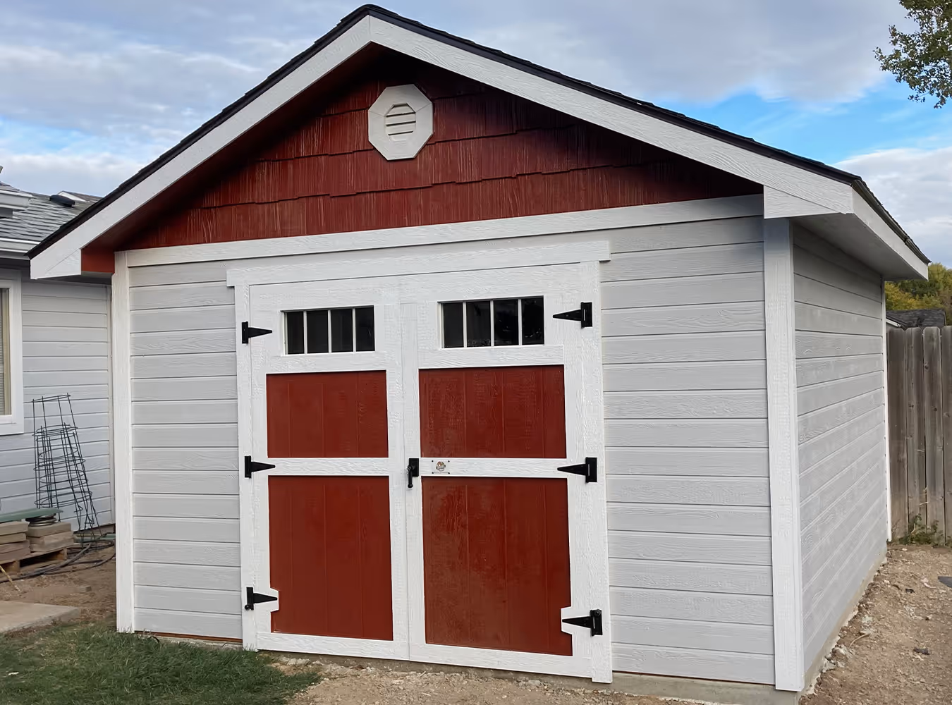 Small light gray shed with a gable roof and double red doors framed in white, featuring small windows at the top.