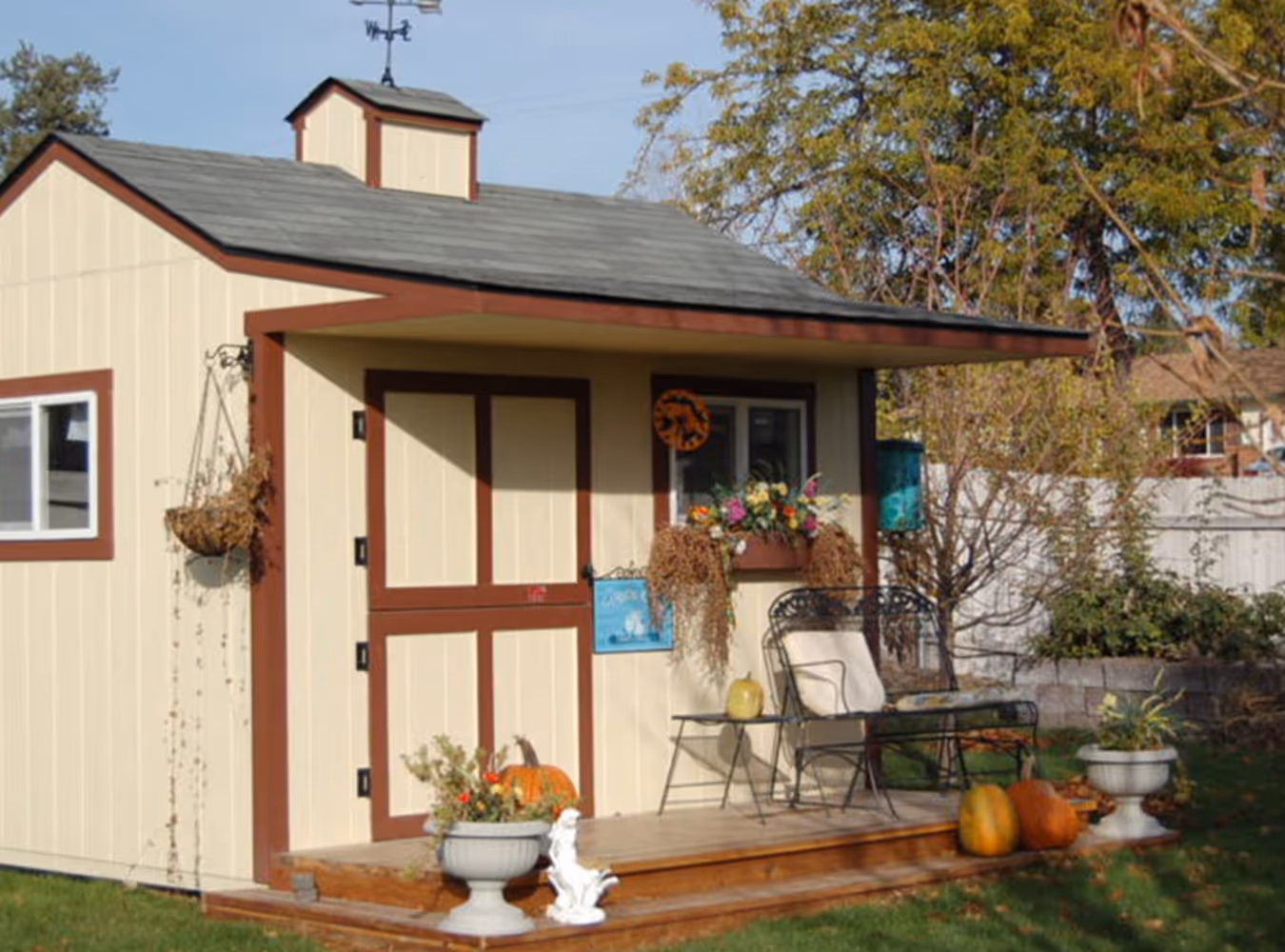 Small beige garden shed with brown trim, a small porch with chairs, pumpkins, flower pots, and dried plants.