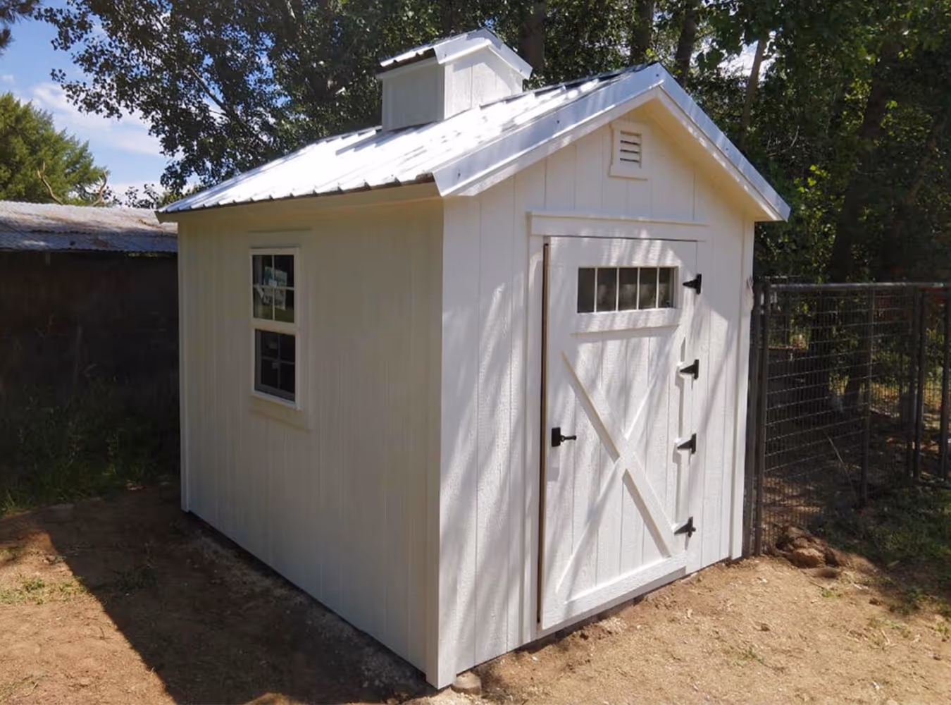 White wooden garden shed with a small window and cross-braced door with black hinges, situated outdoors near trees and a wire fence.