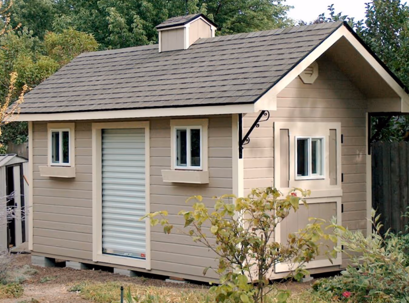 Beige wooden garden shed with gray shingled roof, small windows, and a roll-up door, surrounded by greenery.
