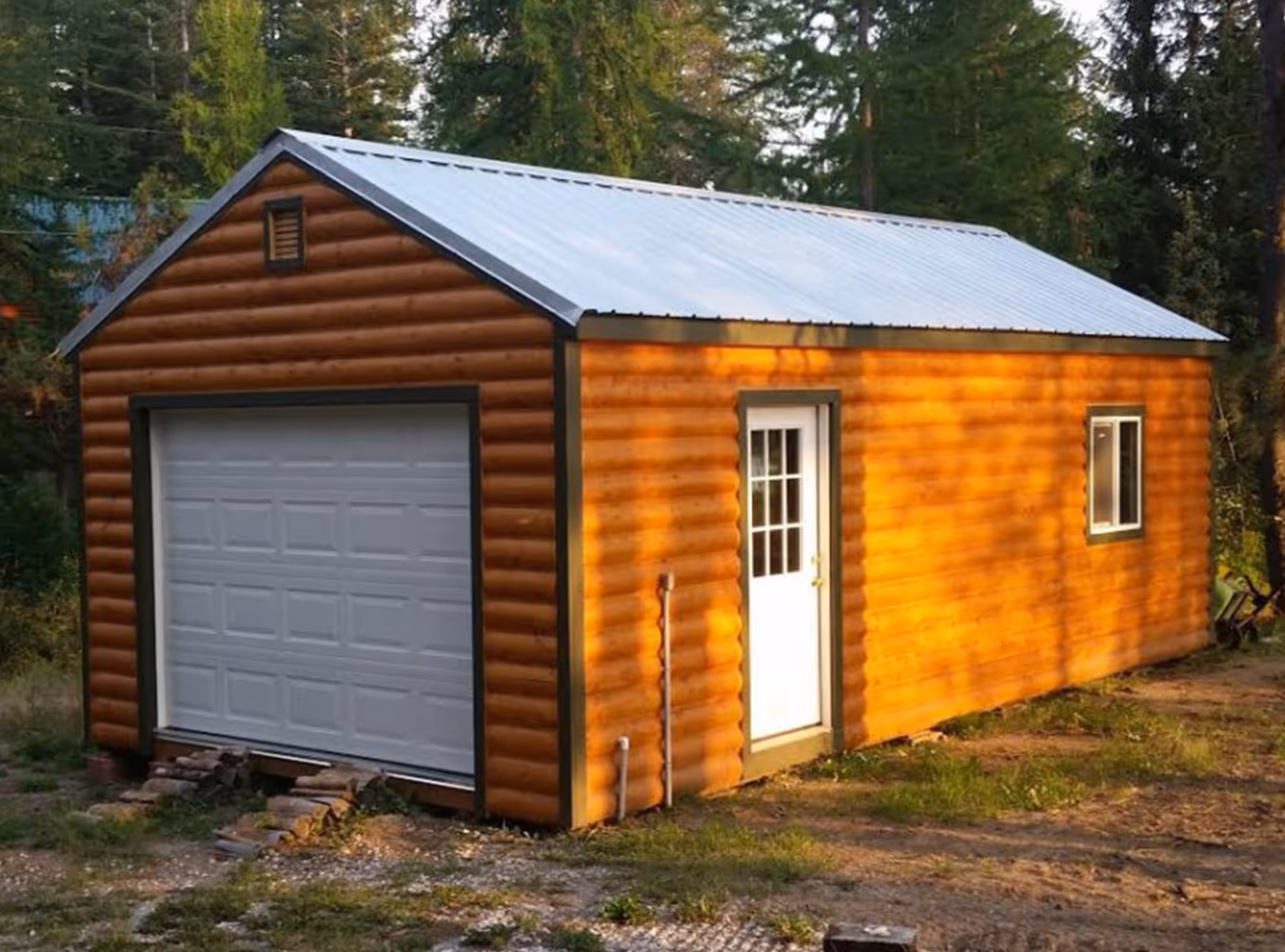 Wooden garage with log-style siding, white garage door, side entrance door, and window, surrounded by trees.