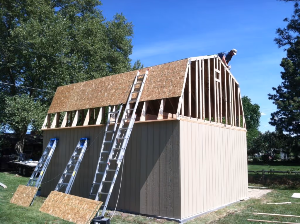 Partially constructed beige barn with a tall gambrel roof under construction on a grassy yard with ladders leaning against it and a worker on the roof.