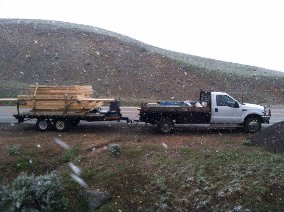 White pickup truck with a flatbed towing a trailer loaded with wooden planks parked beside a road during snowfall.
