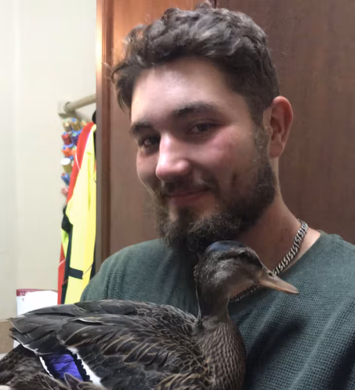 A man with a beard and curly hair holding a brown duck with purple markings indoors.