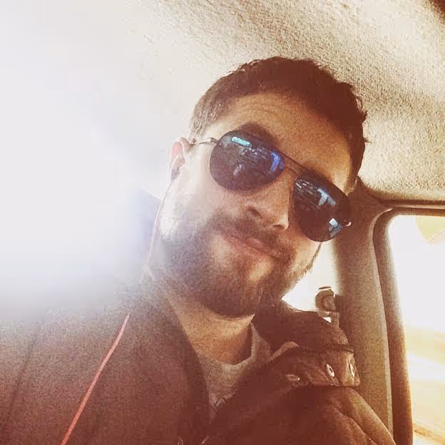 Man with short curly hair and beard wearing dark sunglasses and a jacket inside a vehicle with sunlight coming through the window.