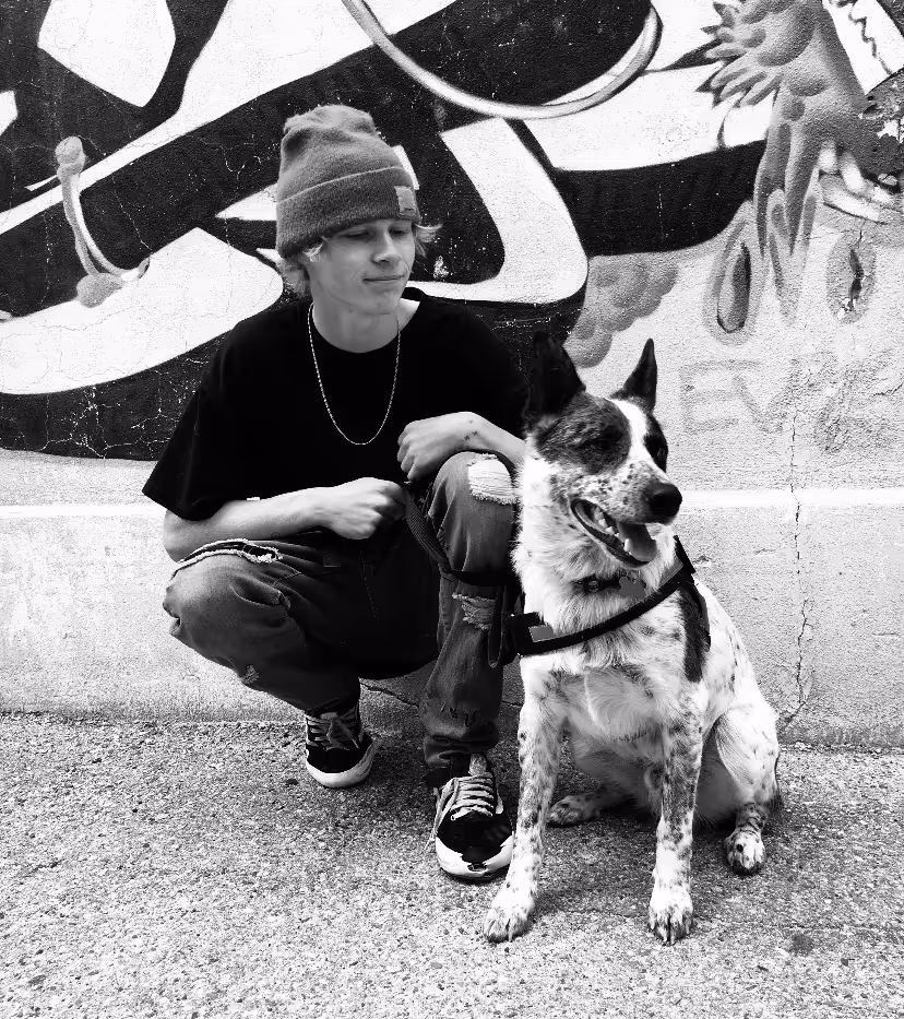 Young person in a beanie and ripped jeans squatting next to a sitting dog with pointed ears against a graffiti wall.
