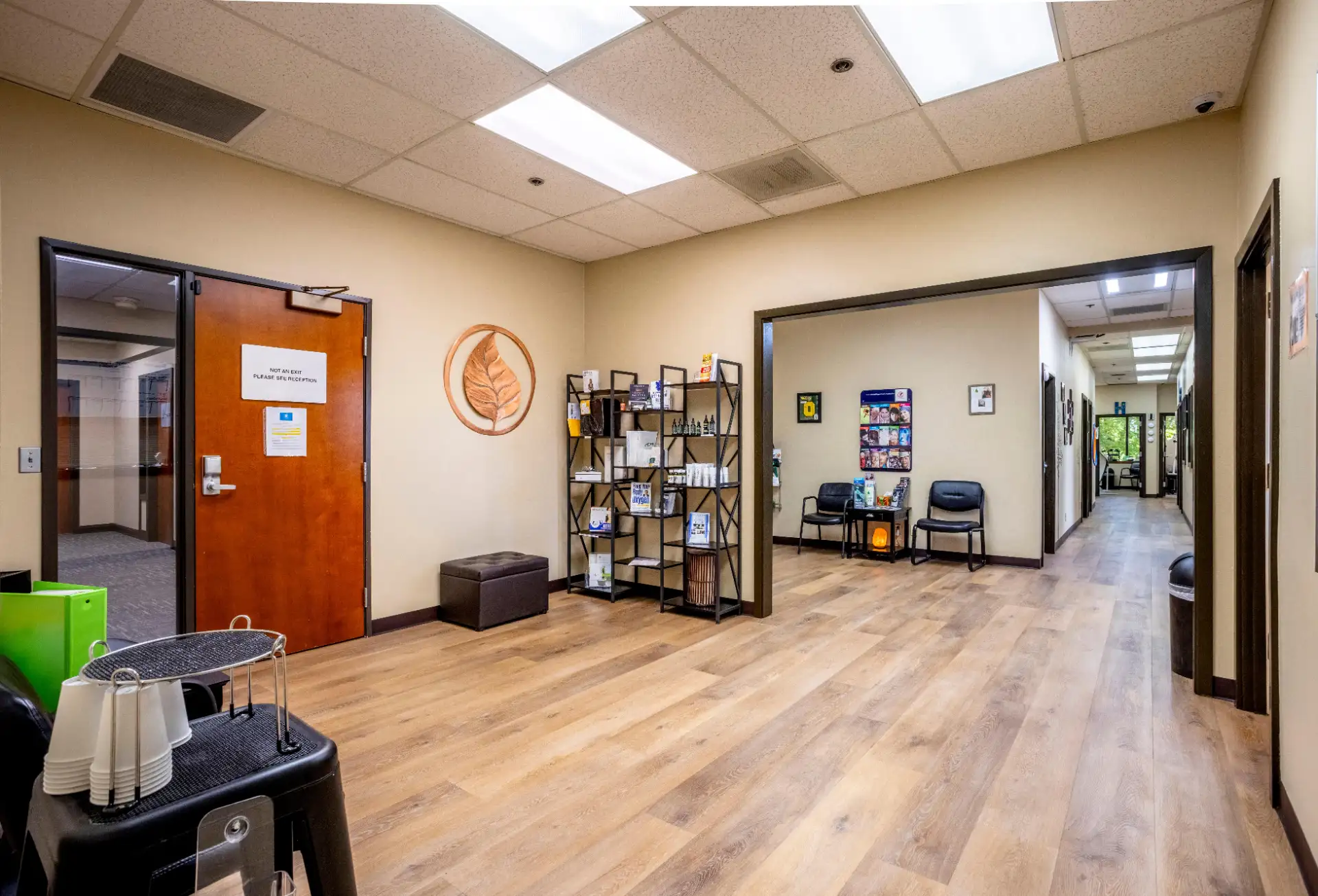 Spacious office lobby with wood floor, beige walls, chairs, shelving with products, and door with exit sign.