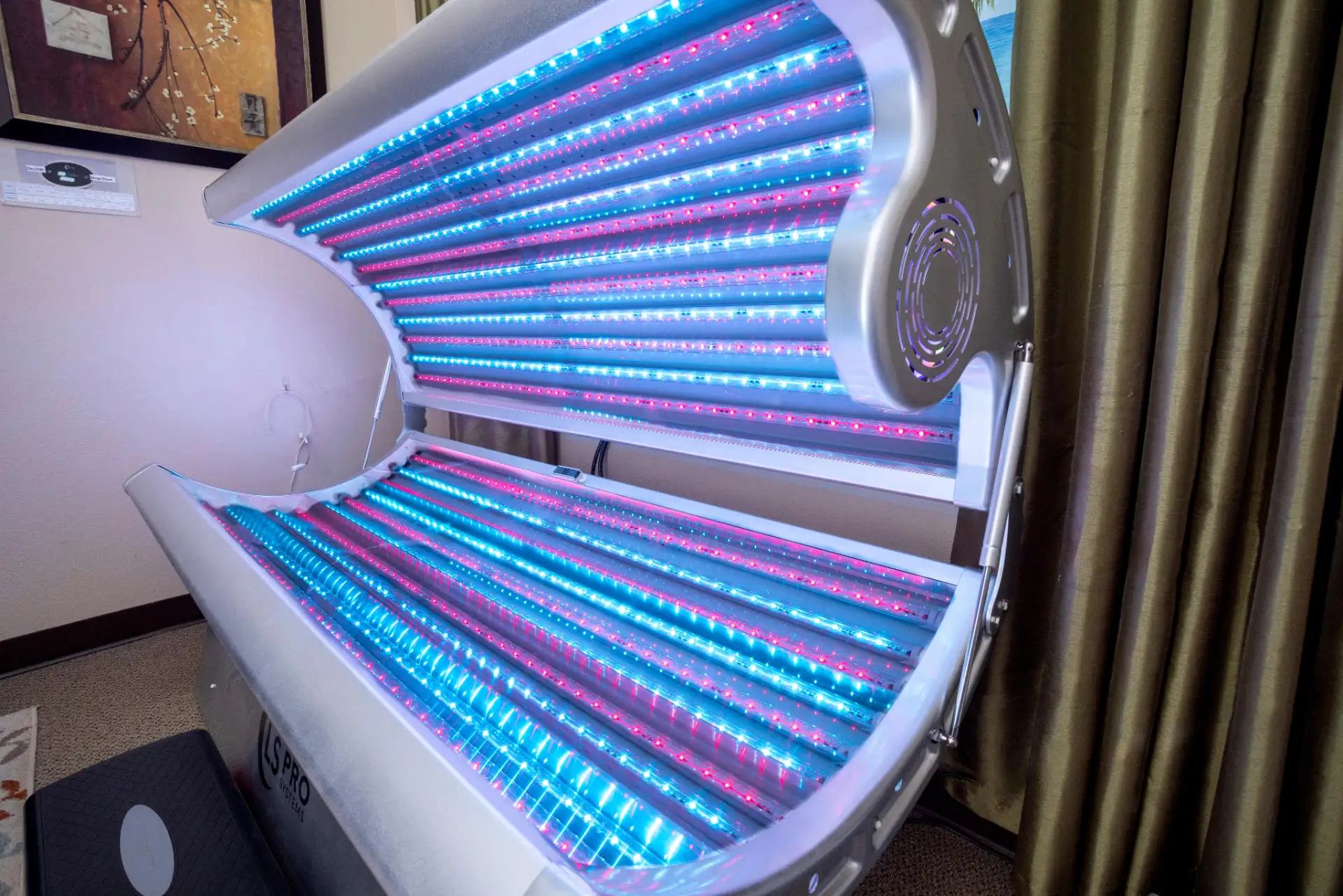 Open tanning bed with blue and red LED lights inside in a room with curtains and a framed picture.