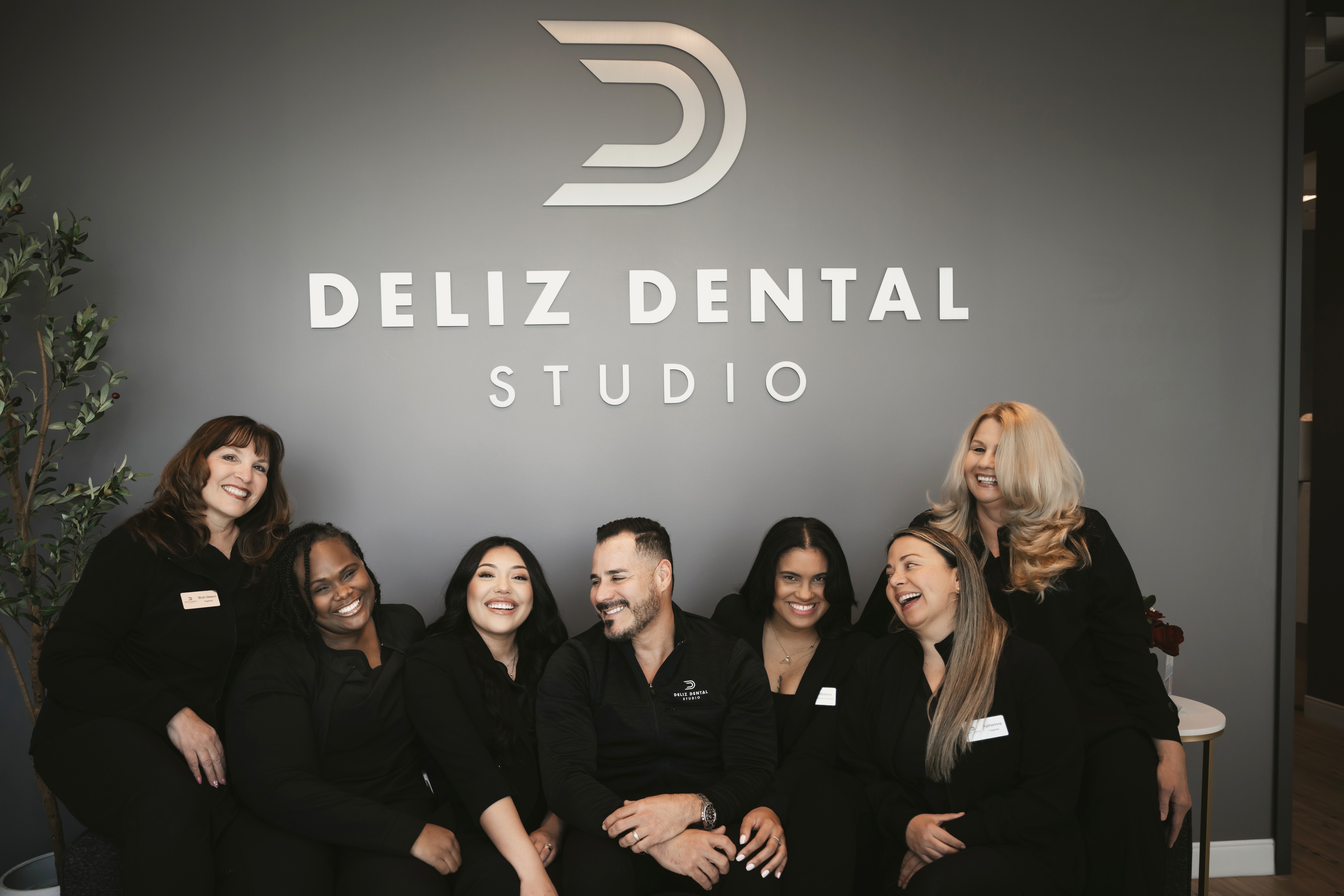 Deliz Dental's Team Posing and Smiling in Front of Their Logo on the Wall of their Office