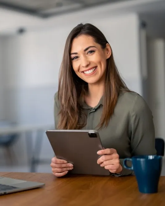 Smiling woman sitting at a desk with a tablet in her hands, a laptop and coffee mug beside her