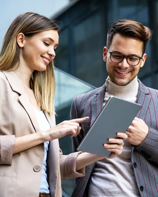 Two smiling colleagues standing outdoors and collaborating on a tablet.