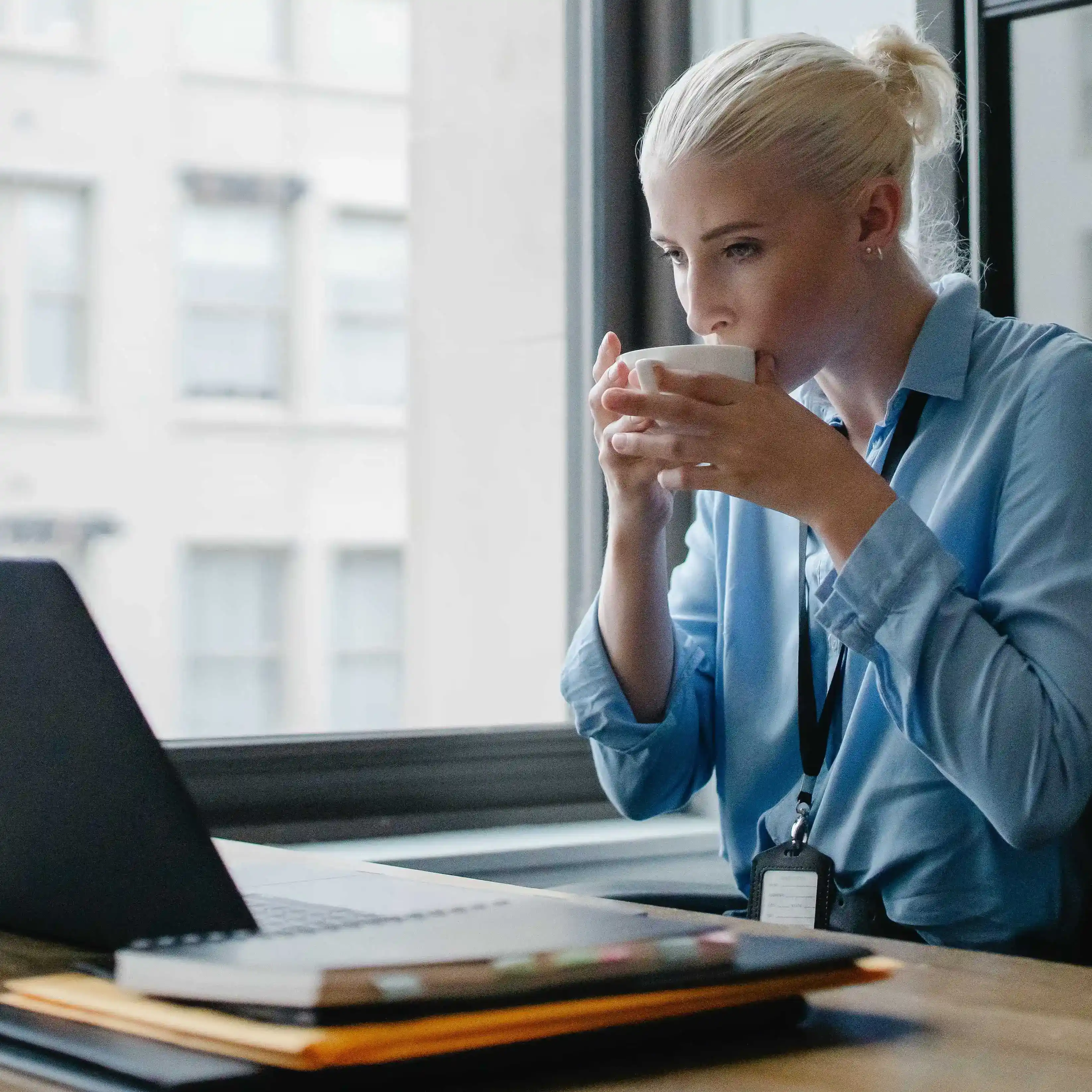 A professional woman sitting at her desk in front of a laptop, near a window, sipping a cup of coffee 