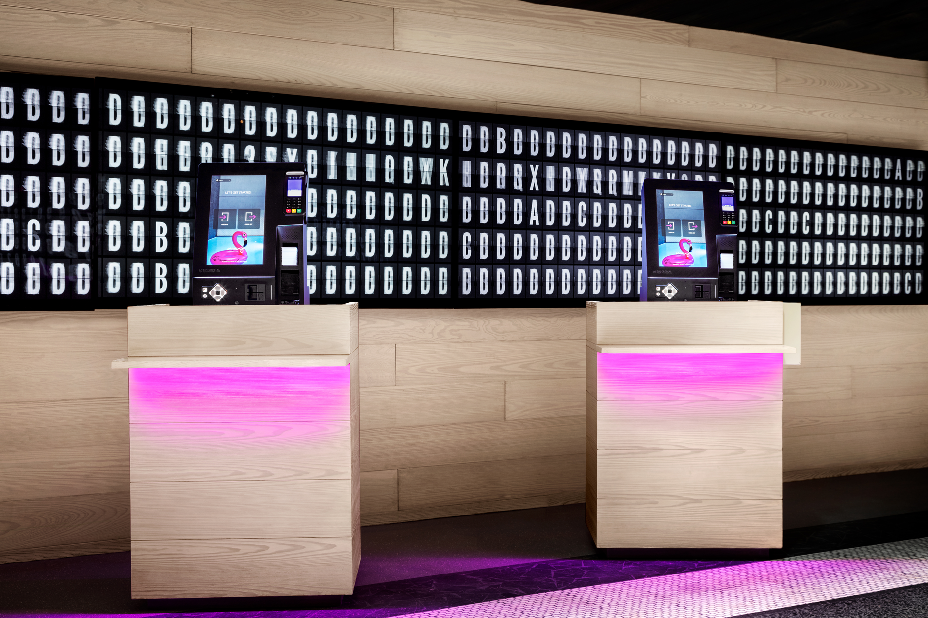 Two self-service check-in kiosks with digital screens and card readers on wooden podiums illuminated with purple lighting, in front of a wall with illuminated lettered panels.